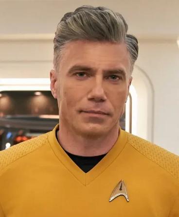 Headshot of Christopher Pike from the shoulders up with a neutral expression wearing a gold colored shirt and insignia badge